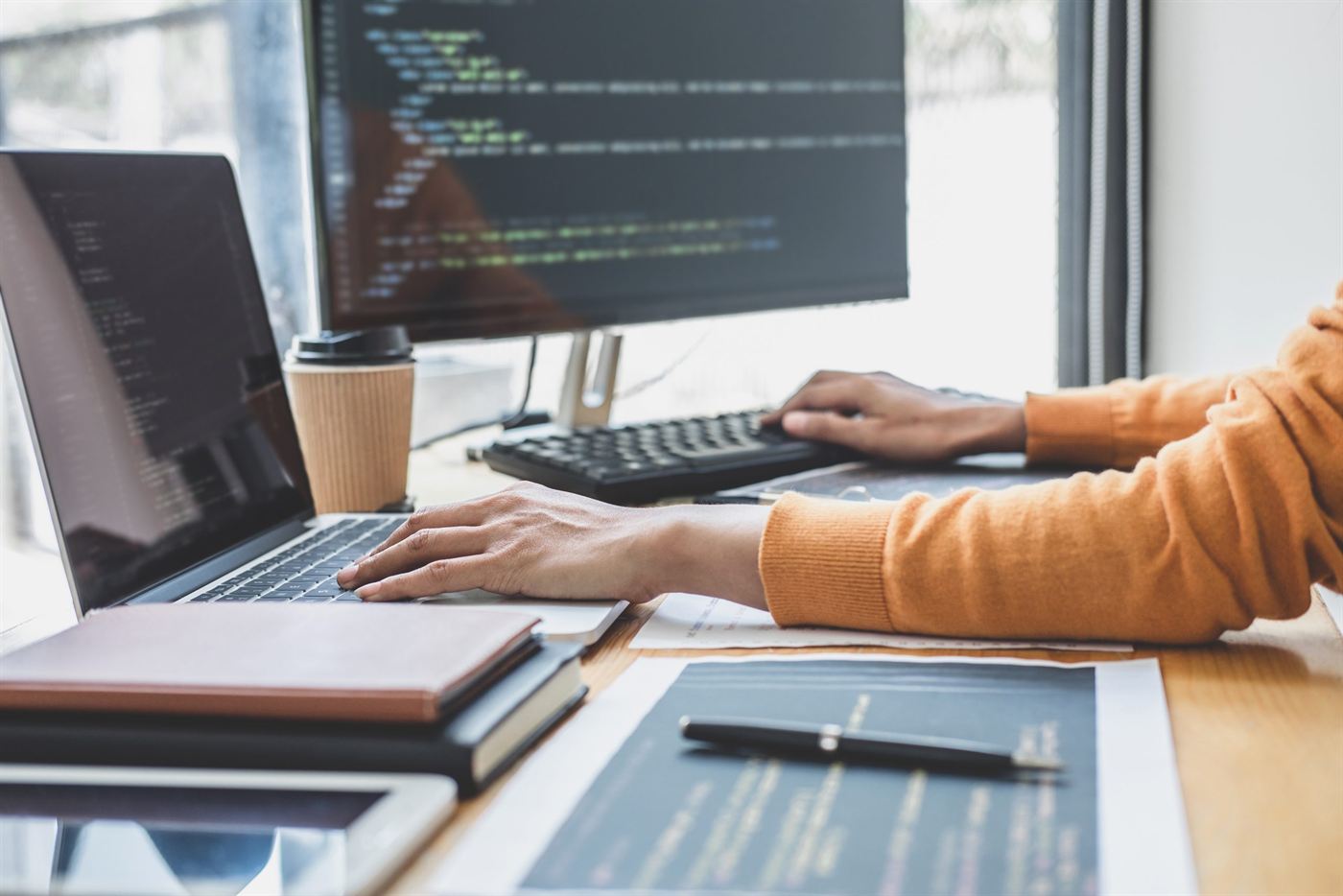 A developer writing code at a desk with a laptop and monitor