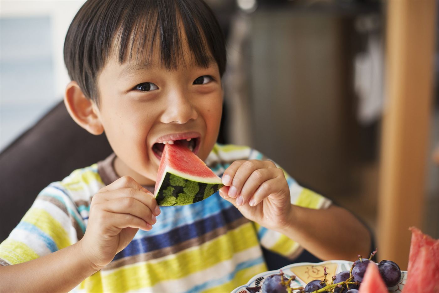 A young boy in a striped shirt smiling as he eats a slice of watermelon